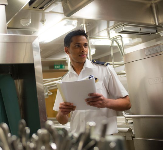 Crew member in uniform holding documents in a cruise ship kitchen | MSC Cruises Crew member in uniform holding documents in a cruise ship kitchen | MSC Cruises