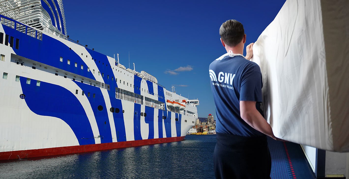 A GNV staff member carrying a mattress onboard a large cruise ship docked at a sunny port | MSC Cruises