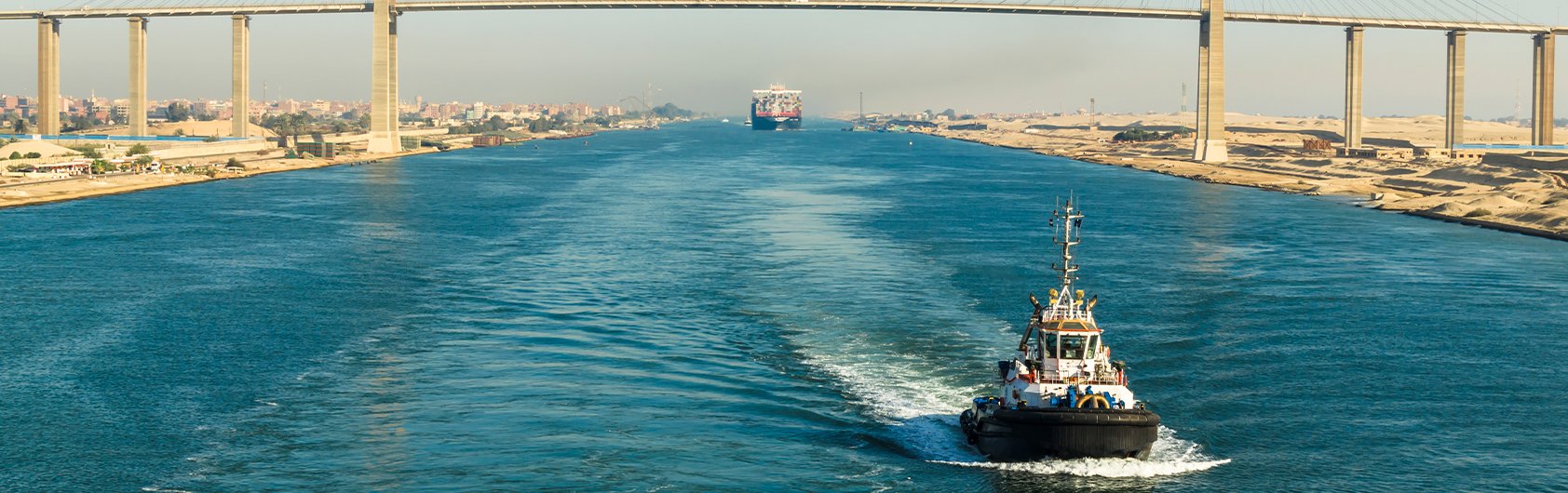 A tugboat navigating the Suez Canal with a bridge and a ship in the background | MSC Cruises